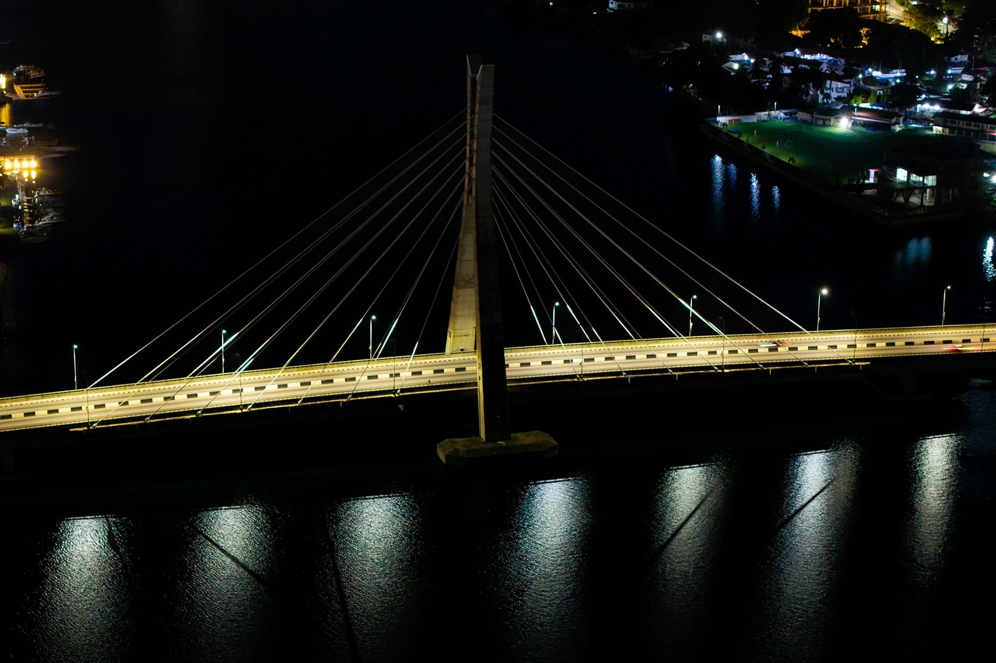 A long-exposure aerial photograph by MO Photos — a cable-stayed bridge at night, lit deck reflecting on dark water.